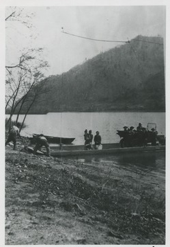 Passengers and an automobile are loaded onto the boat as workers move to send it on its way.