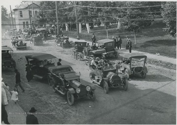 Automobiles line the streets, one of which advertises "Vernon Champe for Congress". A crowd is gathered in the background. 