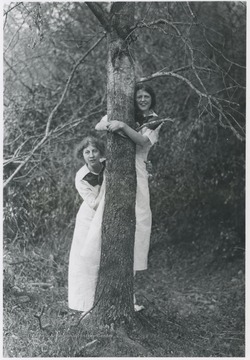 Two unidentified women pose behind a tree. 