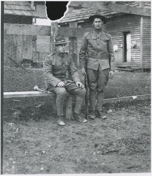 Two men in uniform are pictured by a fence. One sits next to a bugle, the other stands with a rifle. Subjects unidentified. 