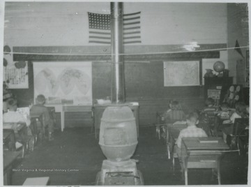 Interior of the school. Children occupy desks facing the chalkboard. Subjects unidentified. 