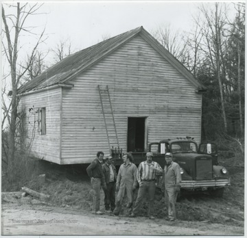 Five unidentified men pose in front of the truck that is hauling the school building. 