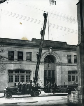 Print number 1266. Firemen pose on the ladder and the truck.  Doc, the Morgantown Fire Department dog, is on the truck beneath the ladder.