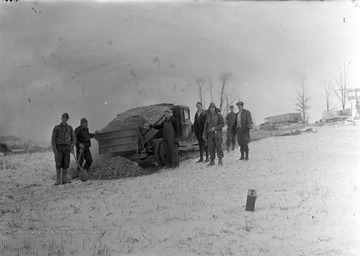 Group of men stand around dump truck on a snowy day, carrying gravel to be used in construction of Franklin Roosevelt's New Deal project.