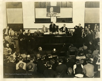 Surrounded by an audience, including local media such as WCHS Radio, with a large photograph of President Franklin Roosevelt hanging over his head, Rush Holt drives home his message.