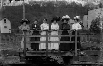 Unidentified group of women, some wearing fashionable hats of the day, lean on the bridge railing. 