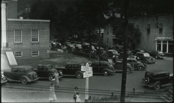 Women are pictured crossing the parking lot from the building, which is pictured on the far left. Subjects unidentified. A sign depicts the direction and distance of Alderson, Lewsiburg, Shady Springs, and Beckley.