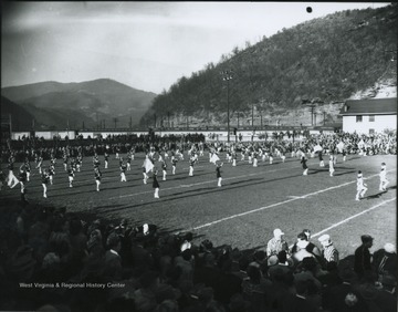The band plays on the field while spectators watch from the bleachers. Referees pictured in conversation on the bottom right.