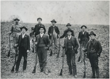 A group of unidentified men pose with their hunting rifles and gear. Two dogs are pictured in the background, likely brought to aid the men in their activities. 