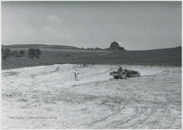 Two unidentified men on the left observe the dug up plot. An unidentified man handles a tractor on the right. 