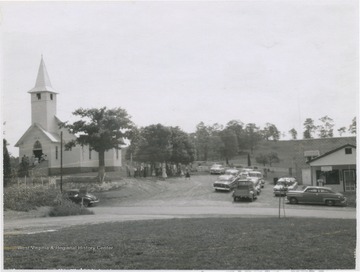 A crowd exits the church, pictured on the left. Cars are parked beside the post office building on the right. 