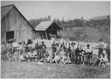 A group of workers pose outside the sawmill. Few men hold onto horses, others hold on to pieces of wood. 