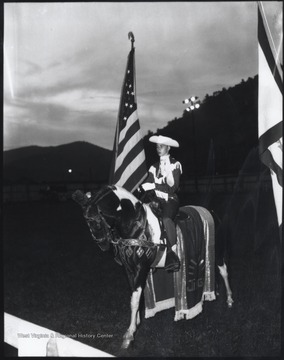 Ralph Damon Williams Jr., son of Dorothy Garten Williams and Ralph Williams Sr., and grandson of Carlos B. Garten who was a sheriff of Summers County, rides with the American flag across the field. The horse is ornamented with elaborate leather straps and an elegant blanket. 