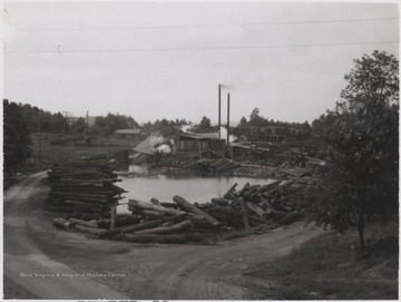 Looking from across the street at the company building and sawmill. Logs surround the pond that sits beside the building. 