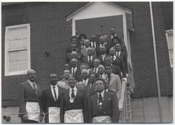 Black Mason members pose on the stairs outside a church. Some of the men are wearing medallions that hold the Free Mason symbol.