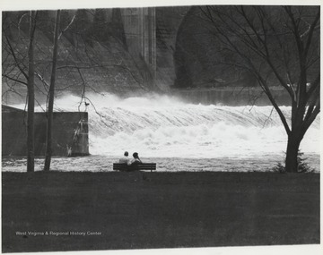 An unidentified couple sits on a bench observing the rushing waters and rapids surging through the dam. 