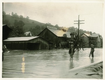 The Leidecker Tool Company building is surrounded by water as some people observe the flood scene from a platform and others wade through the watery street.