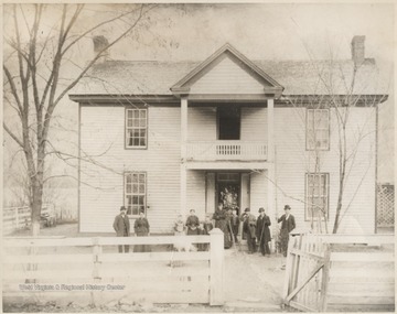 Surveying men and their equipment are pictured outside a large white home. Family members stand closely by. Subjects are unidentified. 
