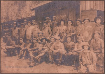 A group of unidentified men pose for a group photo. Some of the men hold hammers. Most of them are dressed in overalls. 