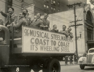 Orchestra leader Tom Whitley is the man wearing glasses on far left pointing forward. The orchestra was the primary performer on "It's Wheeling Steel" radio broadcast. The show was eventually taken to the road, as evident by the "coast to coast" sign. 