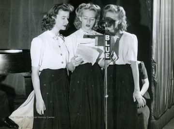 Caption on back of photograph reads: "Here are the Evans Sisters at the microphone, harmonizing a number for listeners to the "Musical Steelmakers". Betty Jane, left, is a receptionist for the company. Margaret June was recently crowned "Miss Steelmaker 1944". Janet Jean, right, age 15, is the youngest member of the trio."