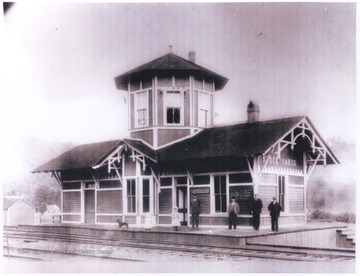 A group of unidentified persons stands outside the building located beside the tracks.