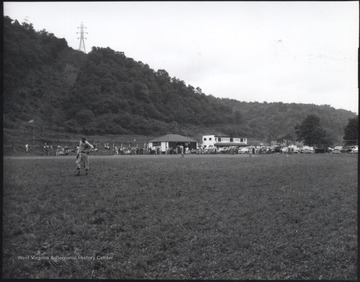 An unidentified baseball player looks at the ground. Behind him, a baseball game ensues. Cars and a crow are lined up along the sidelines. 