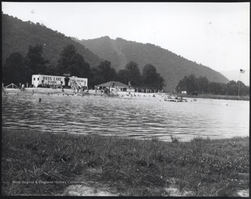 View of the large swimming hole scattered with people. Int he background, the building reads, "Bass Lake Park, Free Picnic Tables".