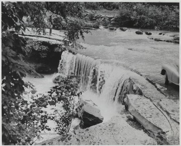 A small waterfall is pictured from the rock above. To the very right, the edge of an automobile is pictured, implying this is a frequently visited spot.