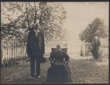 Dennis, standing, and Frances, sitting, are pictured outside on a lawn in front of a white picket fence.