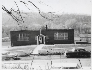 View of the building from across the street. Two automobiles are parked in front of the school yard.