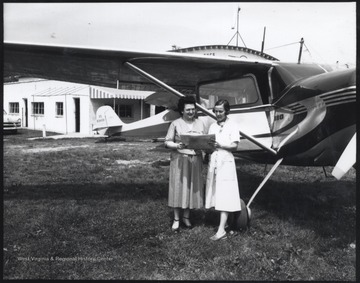 Two unidentified women stand beside an airplane while looking over a map.