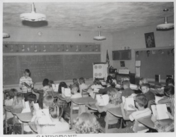 A boy reads in the front of the room while his classmates follow along with their own books. Subjects unidentified. 