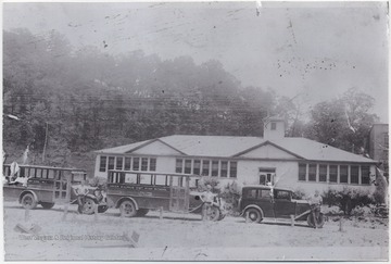 Three unidentified boys stand beside vehicles parked in front of the school building. The school bus in thecenter reads, "Green Sulphur District High School."