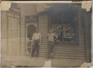 Two unidentified men stand in front of the restaurant's entrance. The windows advertise, "Hot Lunch".