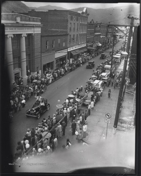 An unidentified man in the leading car sits on top of the seat and waves to either side of the crowded street. Behind him a trail of cars and motorcycles follow.