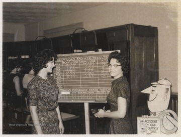 Two unidentified women stand beside a board outlining the week's performance. 