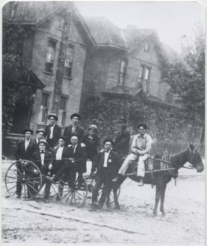 A group of unidentified men sit on top of a horse-drawn wagon outside of the building. 