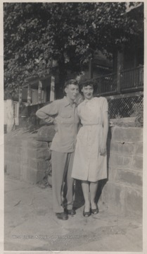 The couple is pictured in front of a stone wall that borders a shaded home. 