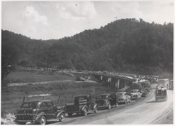 A line of cars are parked along the road while people gather to inspect the new bridge. 