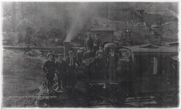 A group of unidentified men pose on top of a transporting train. One of them holds a guitar. 