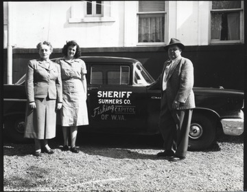 Fox (right) stands beside his police car which claims Summers County as the "fishign capitol of W. Va." The two women on the left are unidentified. 