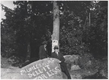 Two unidentified men stand beside a large rock that has painted on it, "Plumley-Hulme: Sell it for less." The Plumley Building was located on the corner of 2nd Avenue and Temple Street, built by William Plumley. 