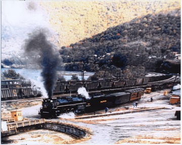 Steam billows from the locomotive as it trails through the railroad yard. 