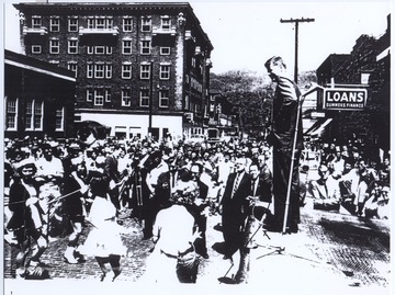 Kennedy smiles at the marching band members to his left. The McCreery Hotel is pictured in the background.