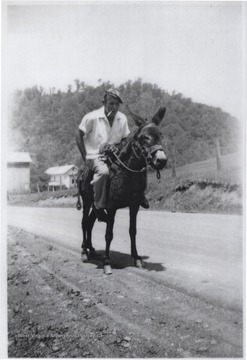 Probably a member of the Shumate family, a man holds himself up on a donkey with a cigarette in his mouth.