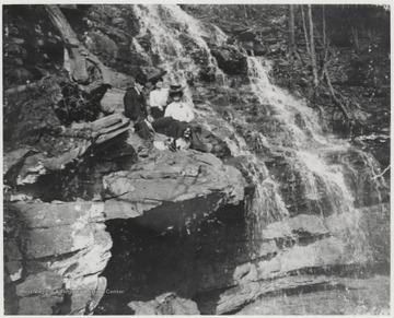 Mr. and Mrs. Robert Murrell, accompanied with an unidentified associate, pose beside the waterfall on a bed of rocks.