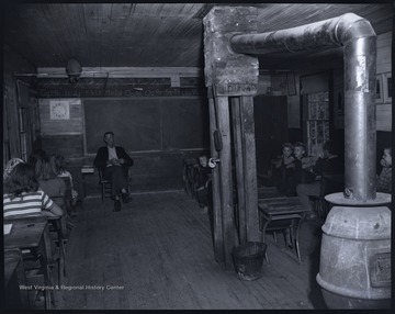 A teacher, likely Charles Saunders, sits in front of the classroom supervising his students. 