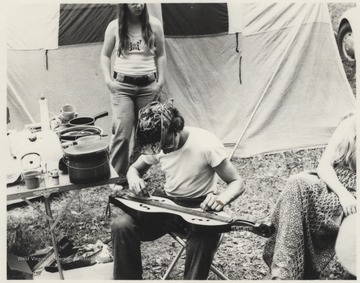 An unidentified man sits beside a table cluttered with pots and pans while plucking at his dulcimer. This photograph is from a series of photos from Appalachian Folk Music Festivals, including the Ivydale and John Henry festivals. 