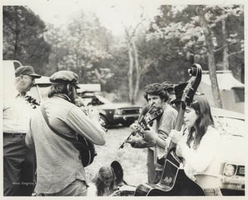 A group of unidentified musicians play beside parked cars. This photograph comes from a series of photos from Appalachian Folk Music Festivals, including the Ivydale and John Henry festivals. 